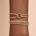 Multicolored braided bracelet with a gold ring on a wrist against a beige background